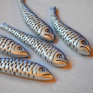 May include: A collection of silver-colored fish sculptures with detailed scale patterns. Each fish has a distinct eye and a slightly downturned mouth, giving them a unique expression. The sculptures are arranged on a neutral-toned surface.