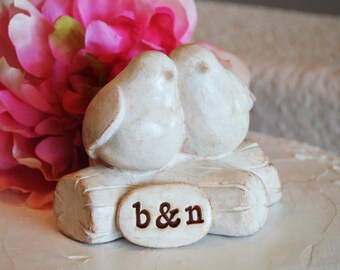 May include: White ceramic wedding cake topper featuring two birds perched on a log with the inscription "b&n" on the front.