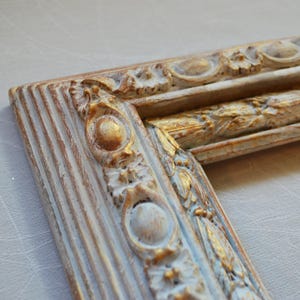 May include: Close-up of a decorative picture frame corner. The frame is painted in a distressed style with cream, gold, and light blue accents. The frame features ornate floral and geometric relief details, adding a vintage aesthetic.