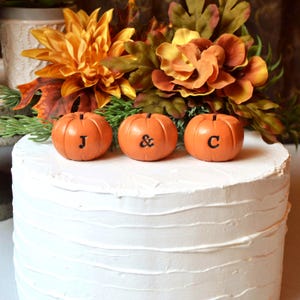 May include: A white frosted cake topped with three orange pumpkin-shaped ornaments. The pumpkins are arranged in a row and feature the black letters J, &, and C engraved on their fronts. Behind the pumpkins are artificial autumn flowers.