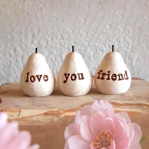 May include: Three off-white, pear-shaped ceramic ornaments with dark brown stems. Each ornament features debossed, dark brown lettering. The first reads love, the second reads you, and the third reads friend, arranged in a horizontal row.