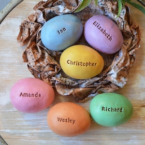 May include: Six colorful, oval-shaped stones with names carved into them: Ian, Elizabeth, Christopher, Amanda, Wesley, and Richard. The stones are pastel shades of blue, lavender, yellow, pink, orange, and green. They rest on a bed of decorative paper.