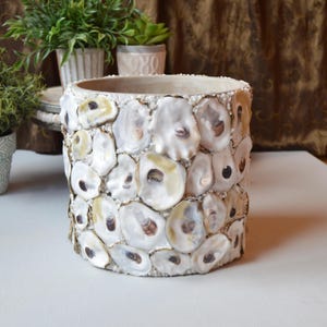 May include: A cylindrical planter covered in overlapping white and cream-colored oyster shells. The exterior surface features a mosaic of natural shell textures with iridescent interiors and dark brown accents, creating a textured, layered appearance.