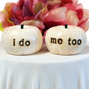 May include: Two white ceramic pumpkins with the words "i do" and "me too" written on them. The pumpkins are sitting on a white tablecloth.
