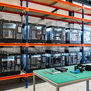 Peut inclure: Une rangée d'imprimantes 3D noires sur des étagères métalliques orange. Les imprimantes ont des enceintes en plastique transparent et sont étiquetées "Raise3D Pro 2 Plus".