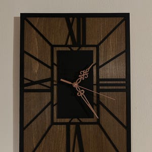 Può includere: Orologio da parete rettangolare con sfondo in legno scuro e sovrapposizione geometrica nera. Il quadrante è nero con lancette color oro rosa e numeri romani. Misura circa 46 cm di altezza e 30 cm di larghezza.