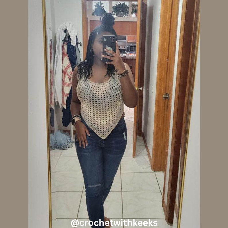 May include: A cream-colored, handmade crochet top with a unique, openwork design. The top has a halter-style neckline and a triangular shape. The model is wearing the top with blue jeans. The text "@crochetwithkeeks" is at the bottom of the image.