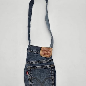 Op de afbeelding: Een blauwe schoudertas gemaakt van gerecyclede spijkerstof, voorzien van een enkele lange schouderband, een achterzak met zichtbare stiksels en een bruin lederachtig merklabel met de tekst Levi Strauss & Co. in rode letters.