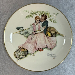 Puede incluir: Plato decorativo con borde dorado, que representa a una pareja, un perro y una cesta de flores. La obra de arte, firmada "Norman Rockwell", muestra una escena romántica sobre un fondo crema. Ideal para decoración.