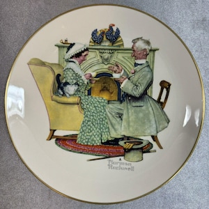 Puede incluir: Plato decorativo con borde dorado que presenta una ilustración de Norman Rockwell. La obra de arte representa a una mujer y un hombre sentados en una habitación con chimenea, un gato y un sombrero de copa. El plato tiene un fondo color crema.