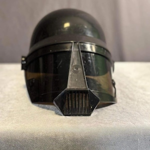 Peut inclure: Casque noir avec un dessus arrondi et une visière. Le casque présente une partie avant angulaire distinctive avec des fentes verticales. Une bande sombre entoure le casque, et le design suggère un thème futuriste ou de science-fiction.