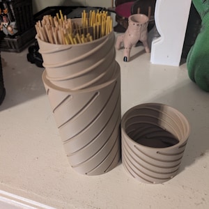 May include: A beige, 3D-printed spaghetti storage container with a spiral design. The container is cylindrical and has a lid. The container is filled with uncooked spaghetti. The lid is off to the side.