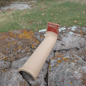 May include: A tan-colored, curved object with a red jar-shaped top. The top has white text and a small, round, dark-colored detail. The object is resting on a gray, textured rock surface with patches of yellow and green.