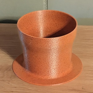 May include: A terracotta-colored planter with a textured surface. The planter has a cylindrical body and a wide, circular base. The top opening is wider than the body. The planter is sitting on a wooden surface.