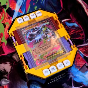 Op de afbeelding: Een gele en zwarte kaarthouder met witte dobbelstenen en een Pokémon-kaart. De kaart zit in een doorzichtige plastic hoes en toont een draakachtig wezen. De dobbelstenen zijn genummerd van 10 tot 100.