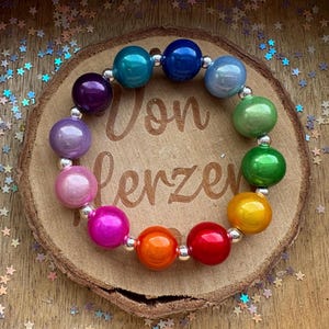 Könnte beinhalten: Ein farbenfrohes Perlenarmband mit einem Regenbogen aus großen, runden Perlen. Die Perlen sind durch silberfarbene Abstandshalter verbunden. Das Armband wird auf einer Holzscheibe mit dem eingravierten Text "Von Herzen" präsentiert.