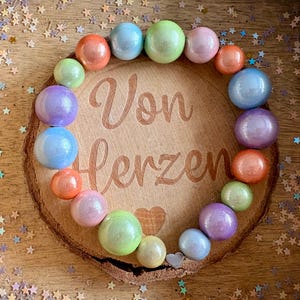 Könnte beinhalten: Eine Holzscheibe mit den Worten "Von Herzen" in Schreibschrift graviert, umgeben von einem Ring aus bunten, irisierenden Kugeln. Die Kugeln sind in den Farbtönen Rosa, Blau, Grün und Orange gehalten.