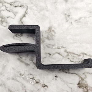 May include: A black desk clamp with a textured surface. The clamp has a C-shaped design, with a top section, a middle section with a slot, and a bottom section with a curved end. It is designed to attach to a desk or table.