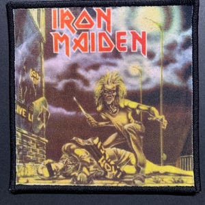 Może przedstawiać: Kwadratowa naszywka z napisem "IRON MAIDEN" w czerwonych literach. Naszywka przedstawia szkieletową postać z długimi włosami, trzymającą broń, na tle sceny cmentarnej. Naszywka ma czarną obwódkę.