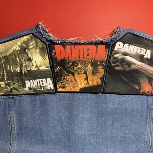 May include: A denim vest with three Pantera band patches. The patches feature album art and the band's name in bold lettering. The background is a solid red.