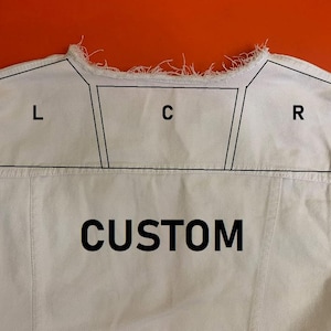 May include: A white denim vest featuring a frayed neckline and armholes. The upper back panel is divided into three sections labeled L, C, and R with black outlines. The word CUSTOM is printed in large black capital letters below these sections.