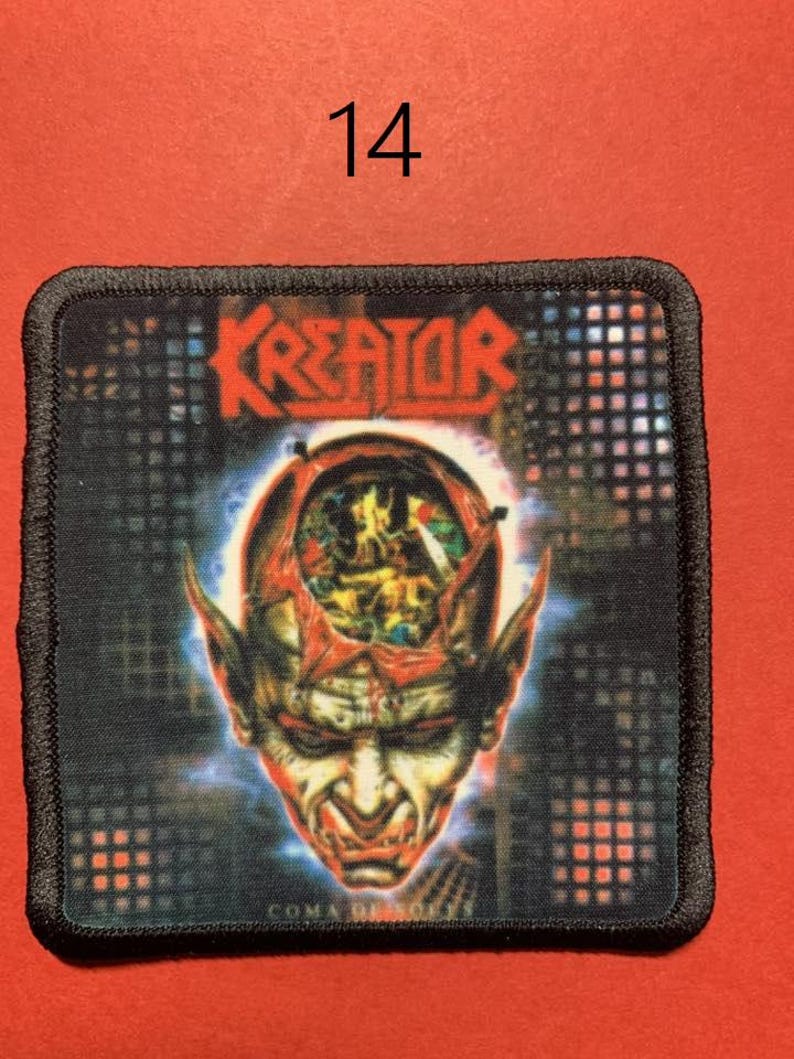 Peut inclure: &Eacute;cusson carr&eacute; avec le mot "KREATOR" en rouge, au-dessus d'une illustration d&eacute;taill&eacute;e d'un visage d&eacute;moniaque avec de la mati&egrave;re c&eacute;r&eacute;brale expos&eacute;e. L'&eacute;cusson a une bordure noire et est pos&eacute; sur un fond rouge. Le chiffre "14" est au-dessus de l'&eacute;cusson.