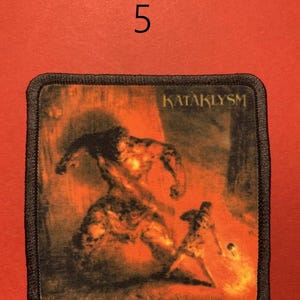 Peut inclure: &Eacute;cusson carr&eacute; &agrave; bordure noire avec une illustration enflamm&eacute;e de deux personnages dans une sc&egrave;ne de bataille. Le mot "KATAKLYSM" est imprim&eacute; en blanc en haut. Le fond est orange flamboyant.