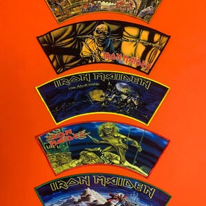 Puede incluir: Cinco parches de tela trapezoidales de Iron Maiden dispuestos verticalmente. Cada parche presenta ilustraciones gráficas distintas y coloridas de la mascota de la banda, Eddie, en varias escenas, con el logotipo de Iron Maiden en letras negritas y estilizadas.