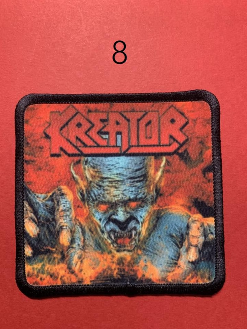 Peut inclure: &Eacute;cusson carr&eacute; avec le mot "KREATOR" en lettres rouges au-dessus d'une figure d&eacute;moniaque aux yeux rouges et aux mains tendues. Le fond est orange et rouge flamboyant. L'&eacute;cusson a une bordure noire et le chiffre "8" est au-dessus.