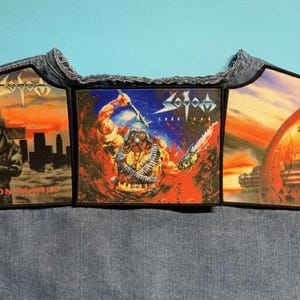 Pode incluir: Três patches com obras de arte de uma banda de metal são exibidos em um colete jeans. Os patches apresentam ilustrações de soldados e cenas apocalípticas. O nome da banda, Sodom, é visível em cada patch. O texto "PERSECUTION MANIA" está em um patch.