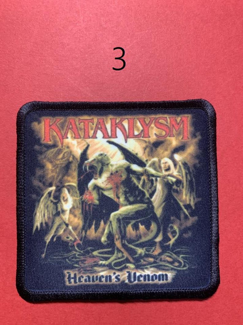 Peut inclure: &Eacute;cusson carr&eacute; avec le mot "KATAKLYSM" en rouge, au-dessus d'une illustration de personnages ail&eacute;s et des mots "Heaven's Venom". L'&eacute;cusson a une bordure noire et est sur un fond rouge. Le chiffre "3" est au-dessus de l'&eacute;cusson.
