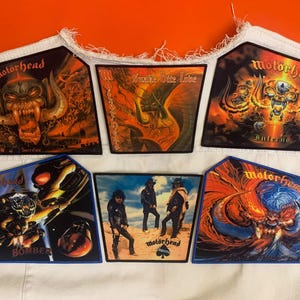 Può includere: Sei toppe in tessuto rettangolari con le copertine degli album dei Motörhead, tra cui Sacrifice, Snake Bite Love, Inferno, Bomber e una foto della band, disposte su un gilet di jeans bianco con bordi sfrangiati su uno sfondo arancione.
