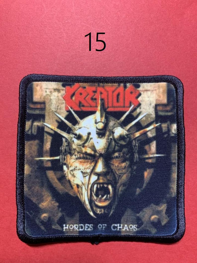 Peut inclure: &Eacute;cusson carr&eacute; &agrave; bordure noire avec le nom du groupe "KREATOR" en rouge et une illustration d'un visage hurlant avec des pointes. Le texte "HORDES OF CHAOS" est en dessous de l'image. Le num&eacute;ro "15" est au-dessus de l'&eacute;cusson.