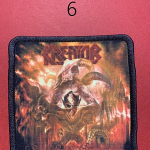 Peut inclure: Un &eacute;cusson carr&eacute; avec une bordure noire pr&eacute;sente une illustration enflamm&eacute;e avec un cr&acirc;ne et des figures d&eacute;moniaques. Le mot "KREATOR" est en haut dans une police stylis&eacute;e. Le chiffre "6" est au-dessus de l'&eacute;cusson.