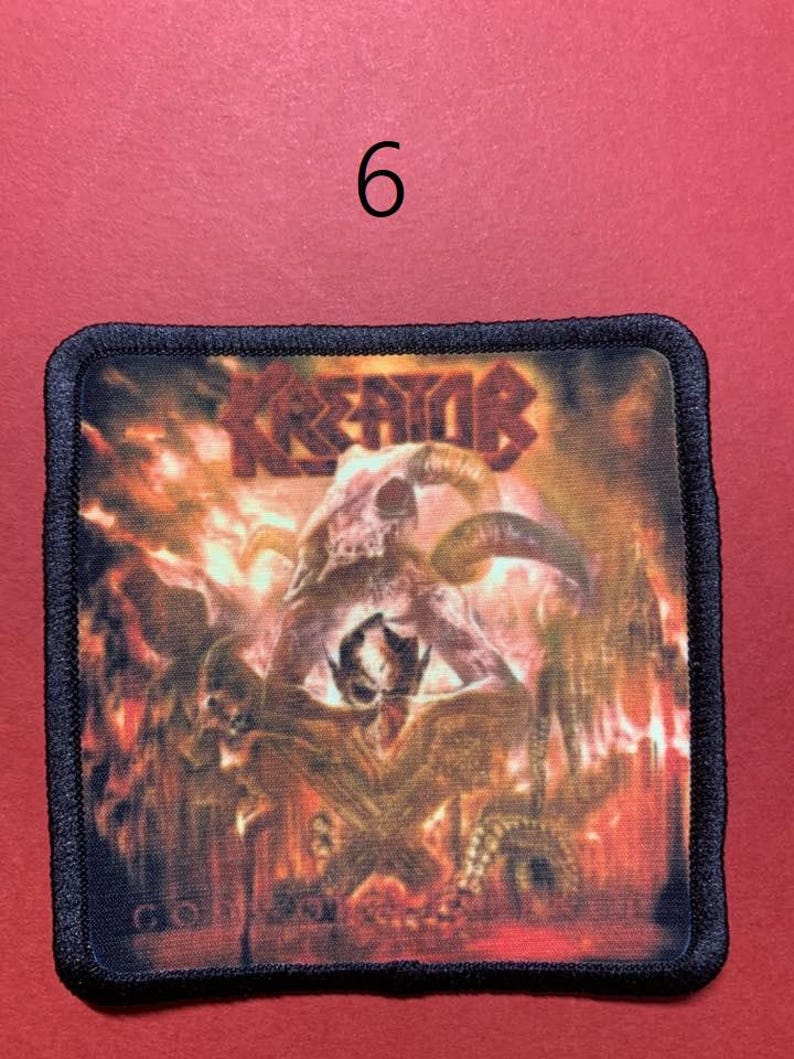 Peut inclure: Un &eacute;cusson carr&eacute; avec une bordure noire pr&eacute;sente une illustration enflamm&eacute;e avec un cr&acirc;ne et des figures d&eacute;moniaques. Le mot "KREATOR" est en haut dans une police stylis&eacute;e. Le chiffre "6" est au-dessus de l'&eacute;cusson.