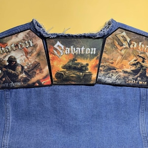 Może przedstawiać: Dżinsowa kamizelka z trzema haftowanymi naszywkami. Każda naszywka przedstawia nazwę zespołu "Sabaton" w stylizowanej czcionce, z motywami wojennymi, w tym żołnierzami i czołgiem. Naszywki są prostokątne z czarną obwódką.