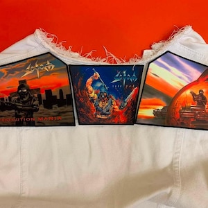 May include: A white denim waistcoat featuring three rectangular patches with graphic illustrations. The left patch shows a soldier with the text Persecution Mania. The centre patch depicts a warrior with the text Sodom. The right patch shows a sunset scene.