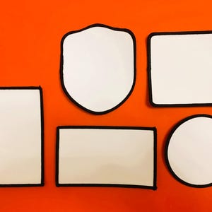 Op de afbeelding: Een verzameling van vijf blanco witte patches met zwarte gestikte randen, bestaande uit een verticale rechthoek, een schildvorm, een vierkant, een horizontale rechthoek en een cirkel, gerangschikt op een effen oranje achtergrond.