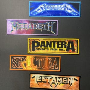 May include: Five rectangular patches displaying heavy metal band logos. The patches feature logos for Metallica, Megadeth, Pantera, Sepultura, and Testament. Each patch has a unique colour scheme, with the band's name clearly visible.