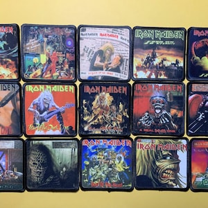 Puede incluir: Una colección de parches de Iron Maiden, cada uno con ilustraciones de álbumes con colores vibrantes y detalles intrincados. Los parches son cuadrados con bordes negros, mostrando varios temas e imágenes asociados con la música de la banda.