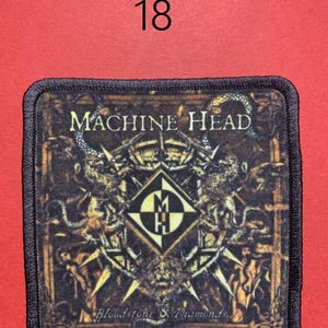 Peut inclure: &Eacute;cusson carr&eacute; avec l'inscription "MACHINE HEAD" en lettres dor&eacute;es. L'&eacute;cusson pr&eacute;sente un motif d&eacute;taill&eacute; avec un embl&egrave;me central et le texte "Bloodstone & Diamonds". Le chiffre "18" est au-dessus de l'&eacute;cusson sur un fond rouge.