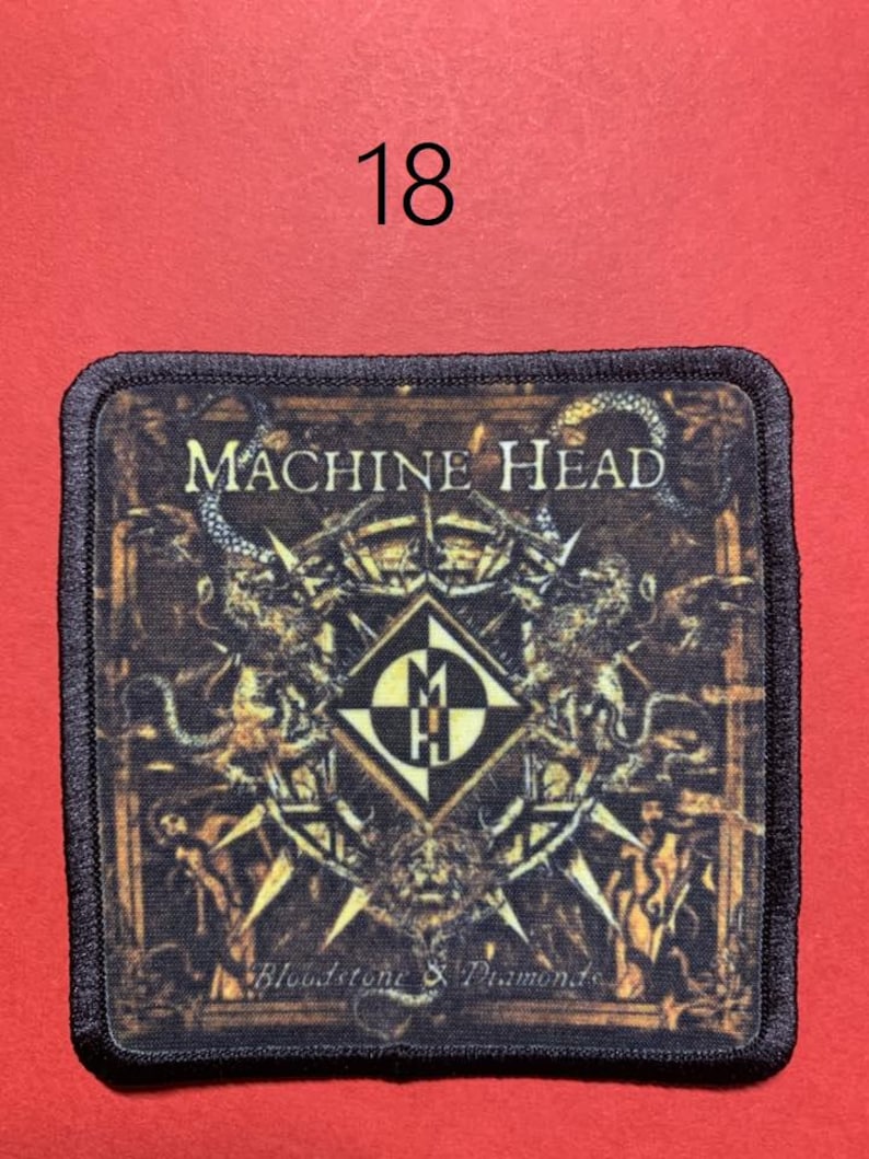 Peut inclure: &Eacute;cusson carr&eacute; avec l'inscription "MACHINE HEAD" en lettres dor&eacute;es. L'&eacute;cusson pr&eacute;sente un motif d&eacute;taill&eacute; avec un embl&egrave;me central et le texte "Bloodstone & Diamonds". Le chiffre "18" est au-dessus de l'&eacute;cusson sur un fond rouge.