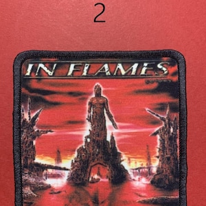 Peut inclure: &Eacute;cusson rectangulaire avec l'inscription "IN FLAMES" dans une police stylis&eacute;e. L'&eacute;cusson pr&eacute;sente un fond rouge et orange avec une figure debout sur une structure. L'&eacute;cusson a une bordure noire et le chiffre "2" est au-dessus.