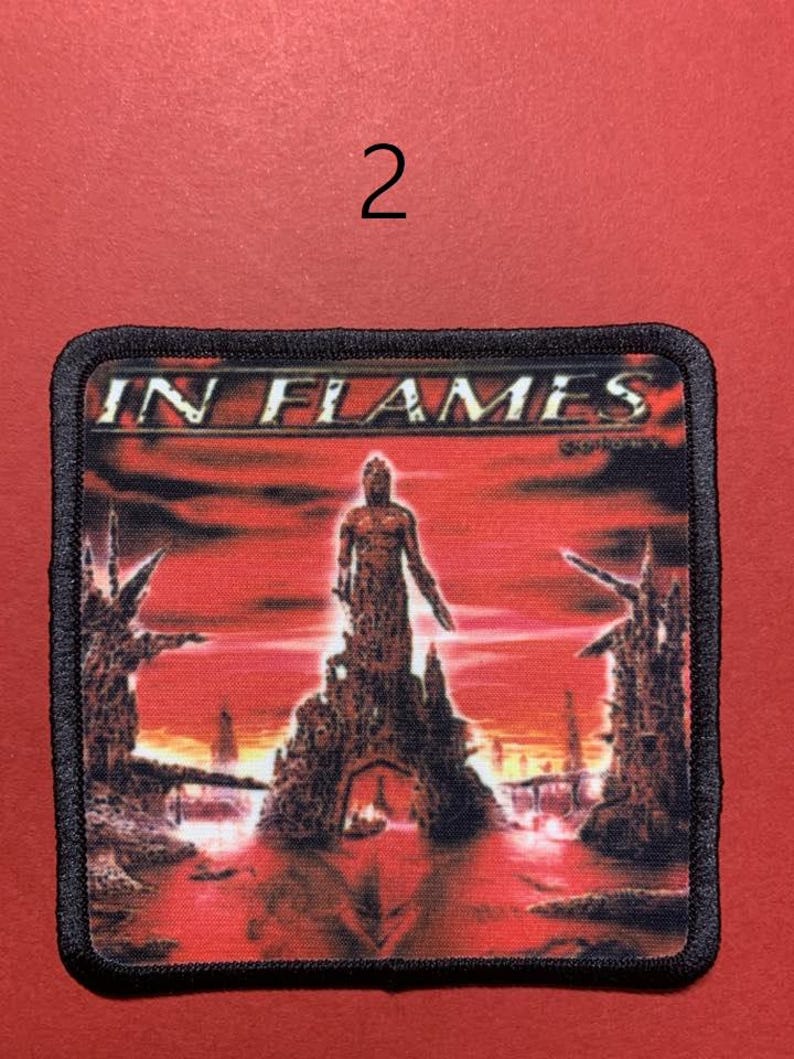 Peut inclure: &Eacute;cusson rectangulaire avec l'inscription "IN FLAMES" dans une police stylis&eacute;e. L'&eacute;cusson pr&eacute;sente un fond rouge et orange avec une figure debout sur une structure. L'&eacute;cusson a une bordure noire et le chiffre "2" est au-dessus.