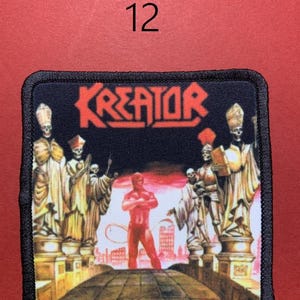 Peut inclure: Un &eacute;cusson carr&eacute; avec le mot "KREATOR" en lettres rouges. L'&eacute;cusson repr&eacute;sente une figure rouge debout sur un pont, avec des figures squelettiques en robes de chaque c&ocirc;t&eacute;. Le chiffre "12" est au-dessus de l'&eacute;cusson.