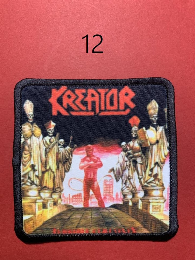 Peut inclure: Un &eacute;cusson carr&eacute; avec le mot "KREATOR" en lettres rouges. L'&eacute;cusson repr&eacute;sente une figure rouge debout sur un pont, avec des figures squelettiques en robes de chaque c&ocirc;t&eacute;. Le chiffre "12" est au-dessus de l'&eacute;cusson.
