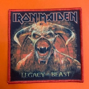 Peut inclure: Un écusson en tissu carré avec une bordure rouge cousue représentant un graphisme de créature à cornes aux dents acérées. Le texte IRON MAIDEN est imprimé en police métallique en haut et LEGACY OF THE BEAST est imprimé en bas.