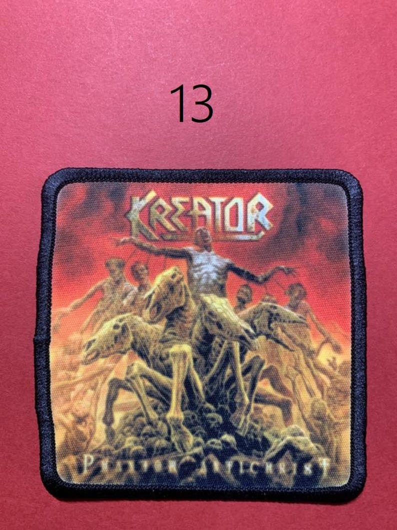 Peut inclure: Un &eacute;cusson carr&eacute; &agrave; bordure noire pr&eacute;sente le nom du groupe "KREATOR" dans une police m&eacute;tallique au-dessus d'une sc&egrave;ne de figures squelettiques et de cavaliers sur fond rouge ardent. Le texte "PRAYER OF ANTECHRIST" est en bas. Le chiffre "13" est au-dessus.
