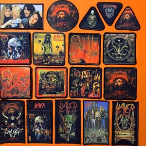 Op de afbeelding: Een verzameling rechthoekige, ronde en driehoekige stoffen patches met diverse Slayer albumhoezen, logo's en bandleden. De patches tonen gedetailleerde, donkere grafische illustraties in rode, zwarte en grijze tinten met zwarte randen.