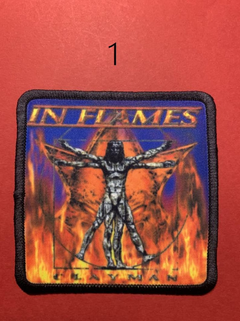 Peut inclure: &Eacute;cusson carr&eacute; avec le texte "IN FLAMES" au-dessus d'une figure de l'Homme de Vitruve dans une &eacute;toile, sur fond de flammes et de ciel bleu. L'&eacute;cusson a une bordure noire et le texte "CLAYMAN" en bas.